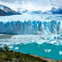 A geleira Perito Moreno, localizada entre os 47º e 51º de latitude sul na Argentina, é uma imponente massa de gelo homenageando Francisco Pascasio Moreno. Estendendo-se do campo de gelo Patagônico Sul ao braço sul do lago Argentino, a geleira possui cinco quilômetros de largura e 60 metros de altura. Considerada uma das maiores reservas de água doce do mundo, está no Parque Nacional Los Glaciares. O fenômeno único da geleira represar as águas do lago resulta em espetaculares desabamentos em intervalos irregulares. Turistas podem observar o evento a uma distância segura, e é possível caminhar sobre a geleira com guias. Descoberta em 1879, foi inicialmente chamada de "Francisco Gormaz" e, posteriormente, "Bismarck". Após disputas territoriais, o tratado de 1899 concedeu soberania argentina, e a geleira foi oficialmente nomeada Glaciar Perito Moreno em homenagem a Francisco Moreno.